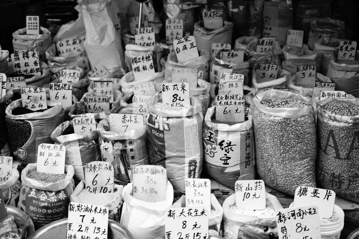 A monochrome market stall display of grains and spices in sacks, each with a small handwritten price tag.