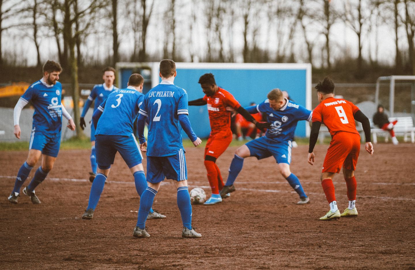 Photograph of an amateur football match on a muddy pitch, players in blue and red kits contesting the ball.