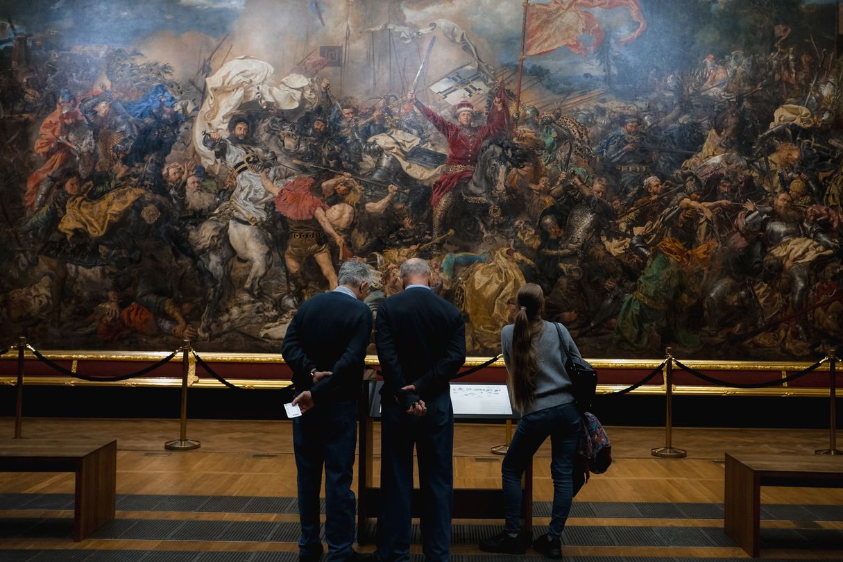 Museum visitors standing in front of a large historical battle painting hung on a gallery wall, most of them looking up at it.