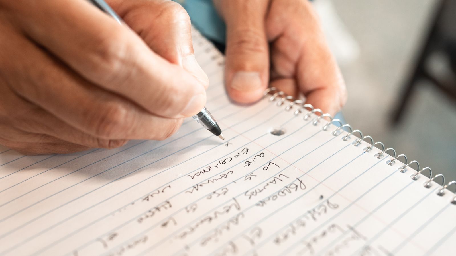 A close-up of hands writing a list in a lined spiral notebook.
