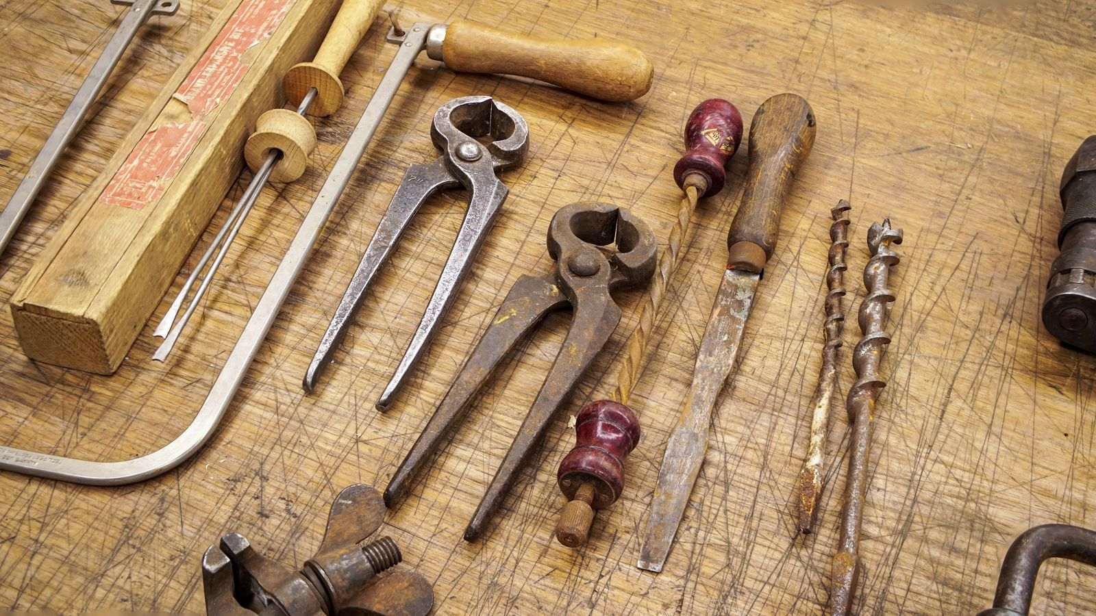 A group of old rusted hand tools laid out on a bare wooden workbench, including a plane, a brace and several chisels.