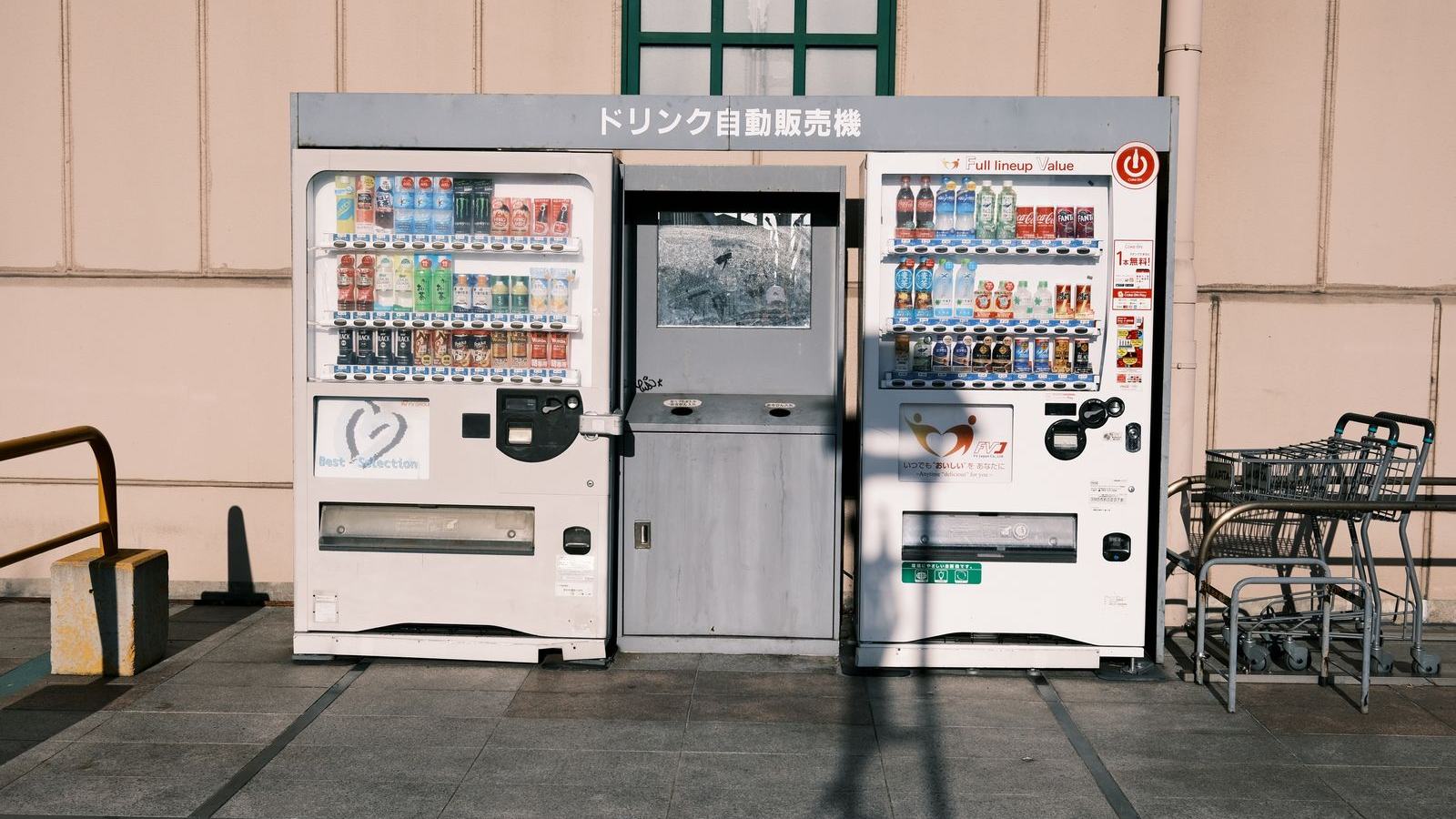 A row of brightly lit drinks vending machines standing on a city street at dusk, their fronts filled with rows of identical bottles and cans.
