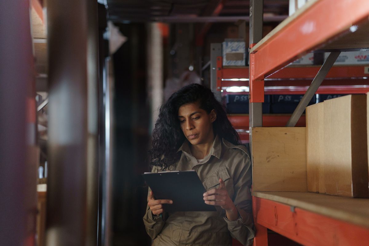 A woman in a warehouse holding a clipboard and looking at stock, taking inventory.