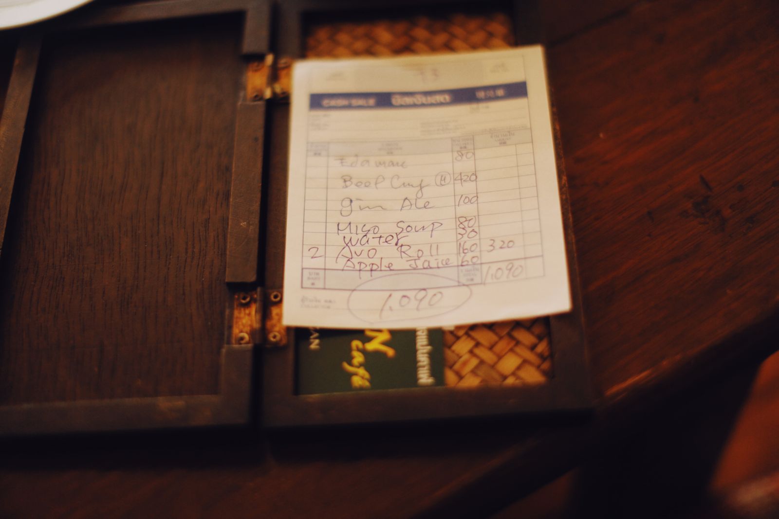 A handwritten cafe bill resting in a hinged wooden bill folder on a dark table, each item listed on its own line with a price against it, and the total circled in pen at the foot.