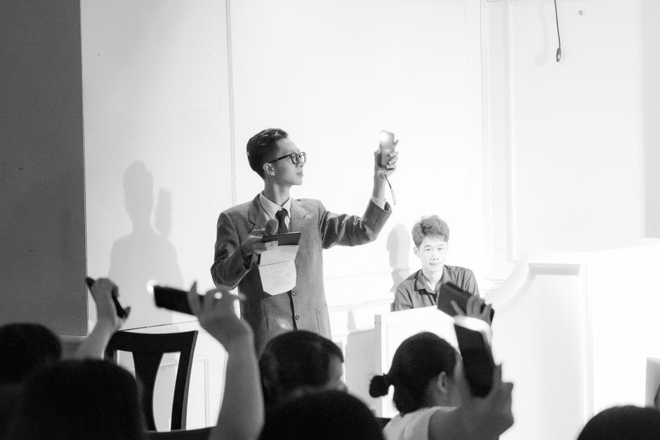 A black and white photograph of a man in a suit and glasses standing on a platform, papers in one hand and a small gavel raised in the other, with an audience in the foreground holding phones up towards him.