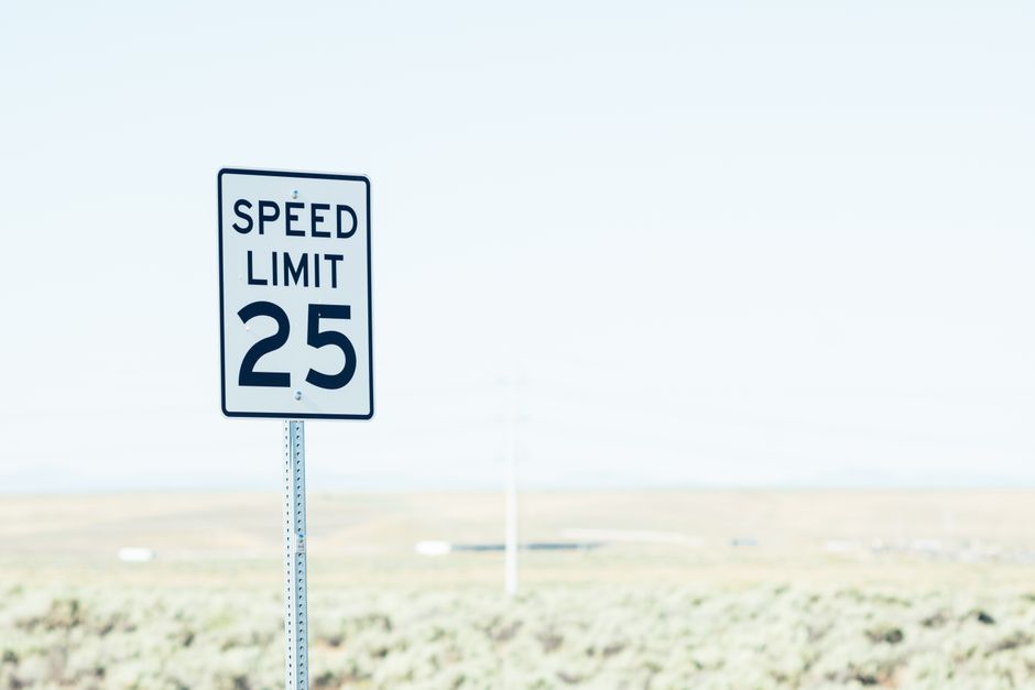 A white American speed limit sign reading twenty five, mounted on a metal post against a pale sky, with dry scrubland and a distant utility pole behind it.