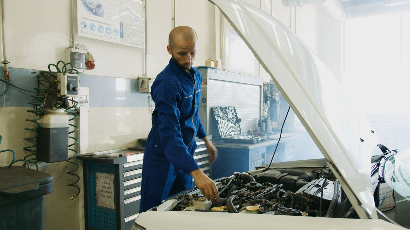 A mechanic in a blue uniform leaning over an open car bonnet, inspecting the engine in a workshop.