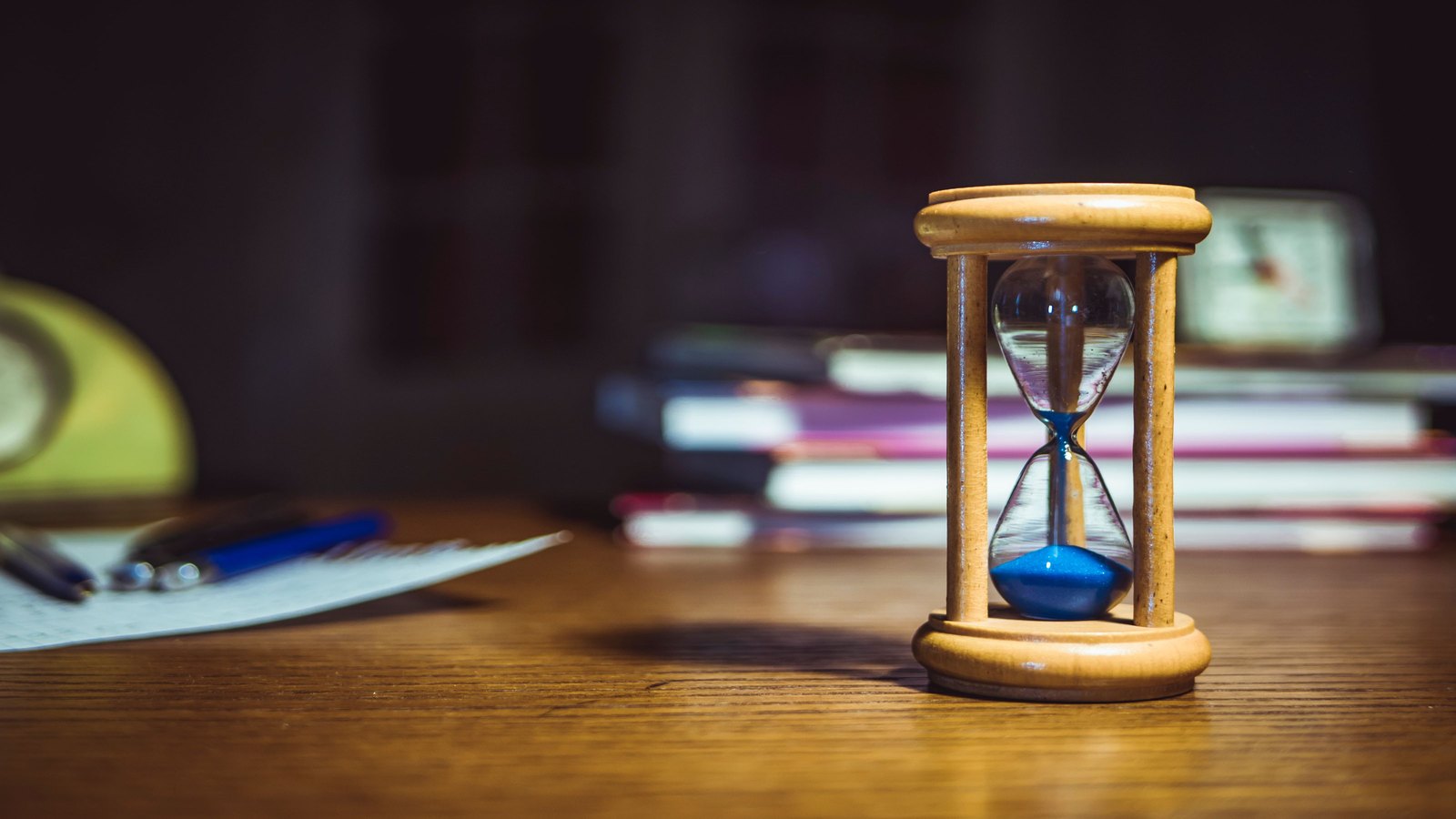 A wooden hourglass standing on a desk beside books and pens, most of its sand still in the upper bulb.