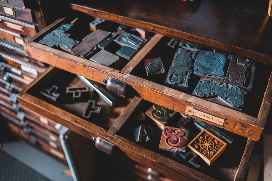 Wooden cabinet drawers in a print workshop, photographed from above, filled with metal letterpress type and assorted printing accessories.