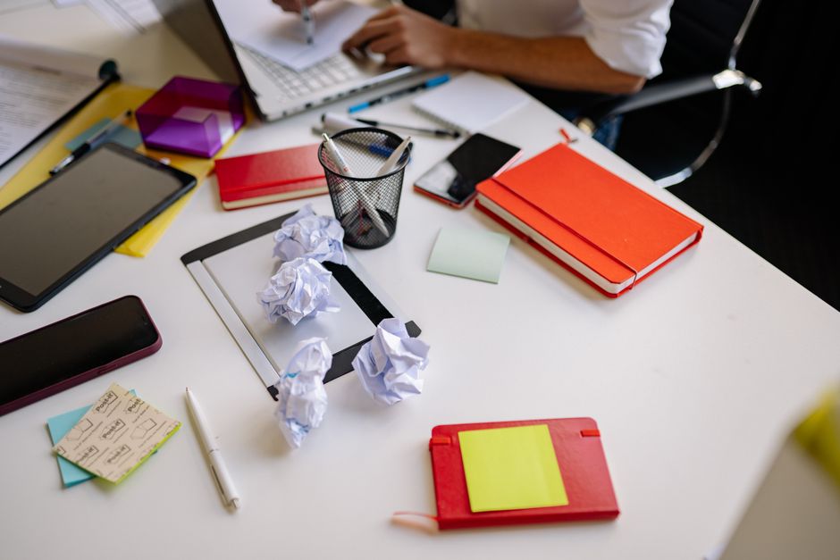 A cluttered office desk covered in crumpled sheets of paper, open notebooks and loose stationery.