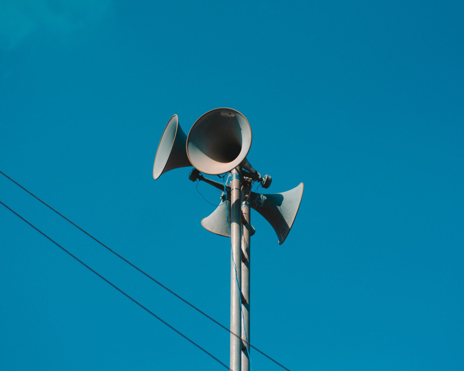 Four grey horn loudspeakers clamped to the top of a metal pole against a clear deep blue sky, each pointing a different way, with overhead cables crossing the lower left of the frame.