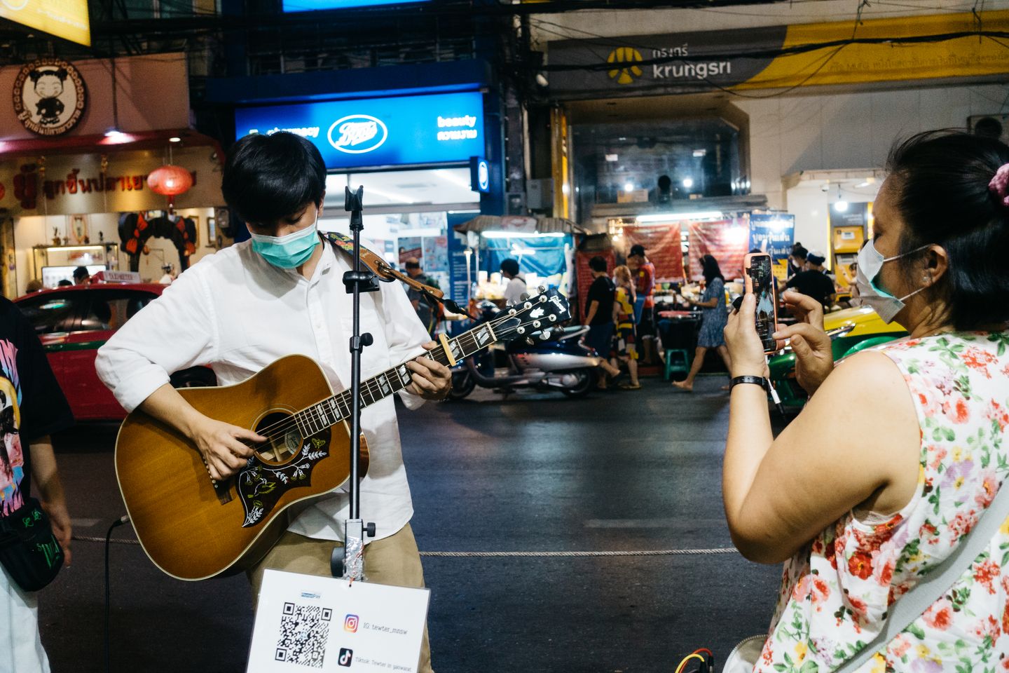 A masked street musician playing an acoustic guitar at night on a busy shop-lined street, a printed QR code sign carrying Instagram and TikTok handles taped below his microphone stand, while a woman in a floral dress films him on her phone.