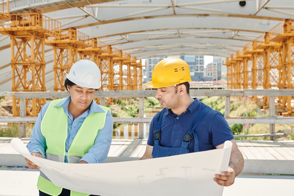 Two people in high-visibility jackets and hard hats standing on a construction site, reading a large blueprint together.
