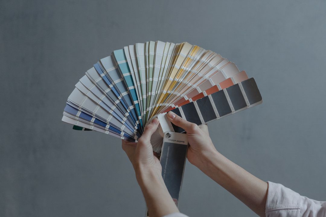 A pair of hands holding an open fan of paper paint swatches, spread wide against a plain grey wall, running through blues, greens, yellows, terracotta and charcoal.