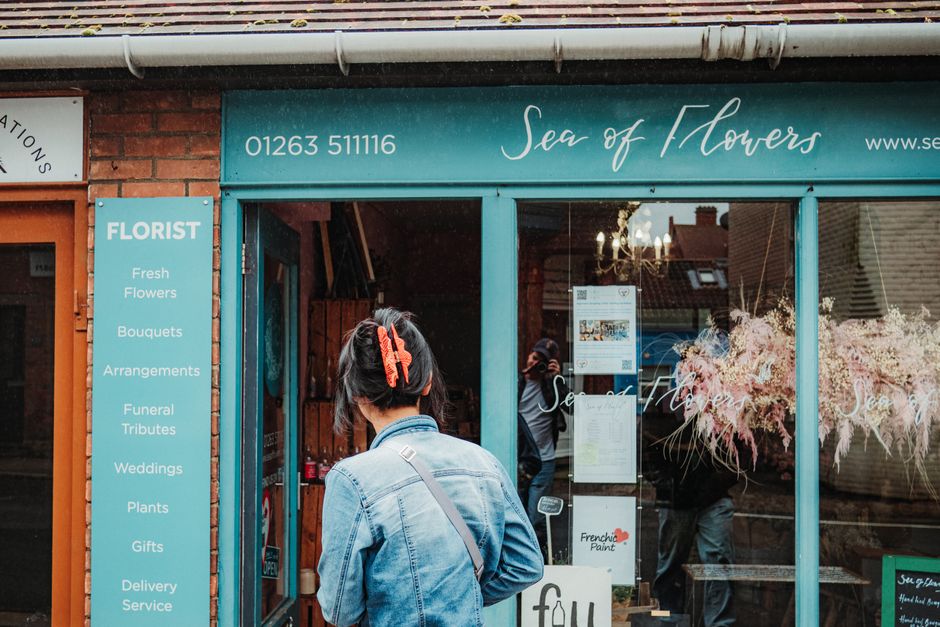 A woman looking into an independent florist: a teal shopfront carrying the shop name in painted script, a phone number, and a painted panel listing what the shop does.