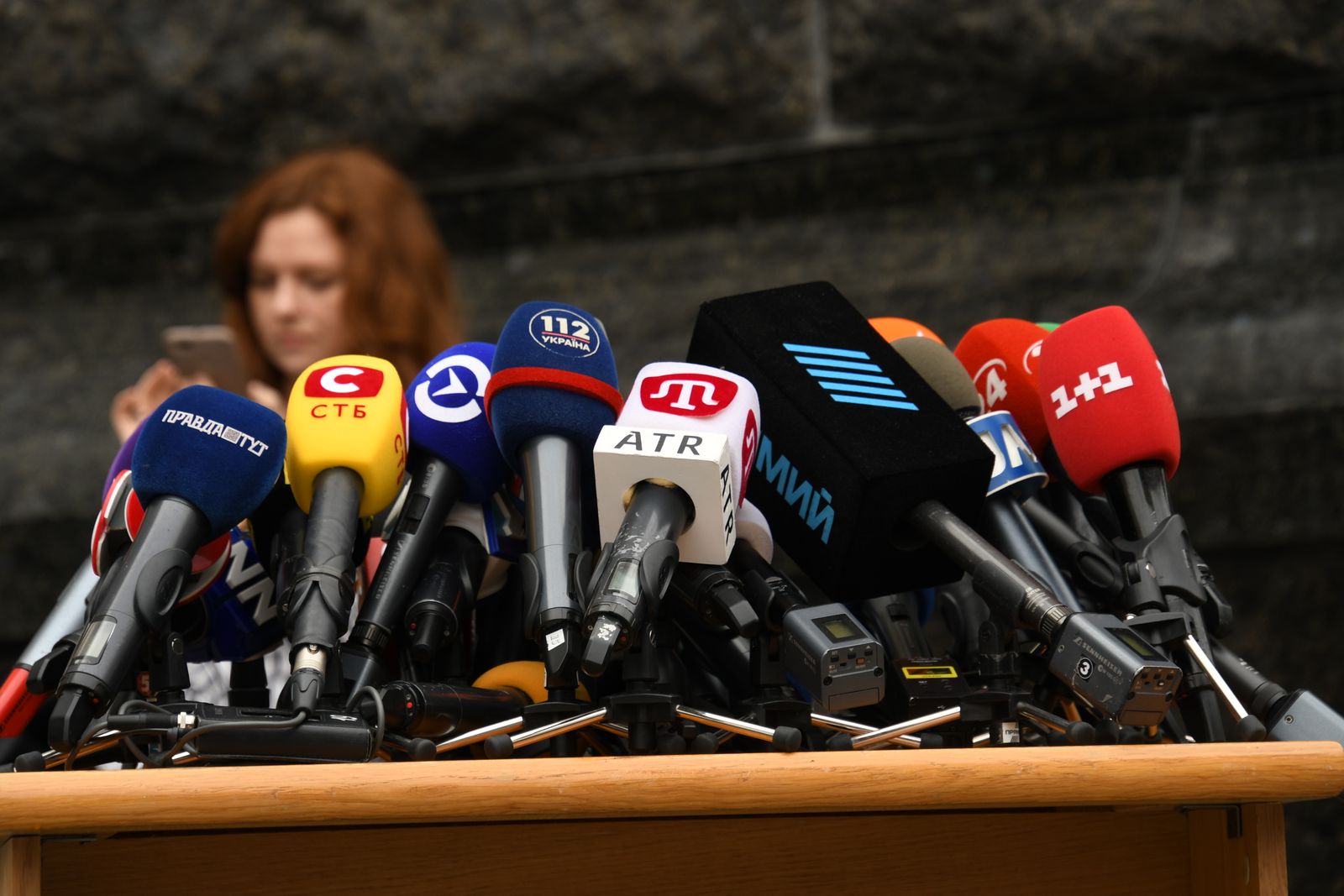 A dense cluster of broadcast microphones from several different organisations, branded with their station logos, standing together on a plain wooden table at a press conference.