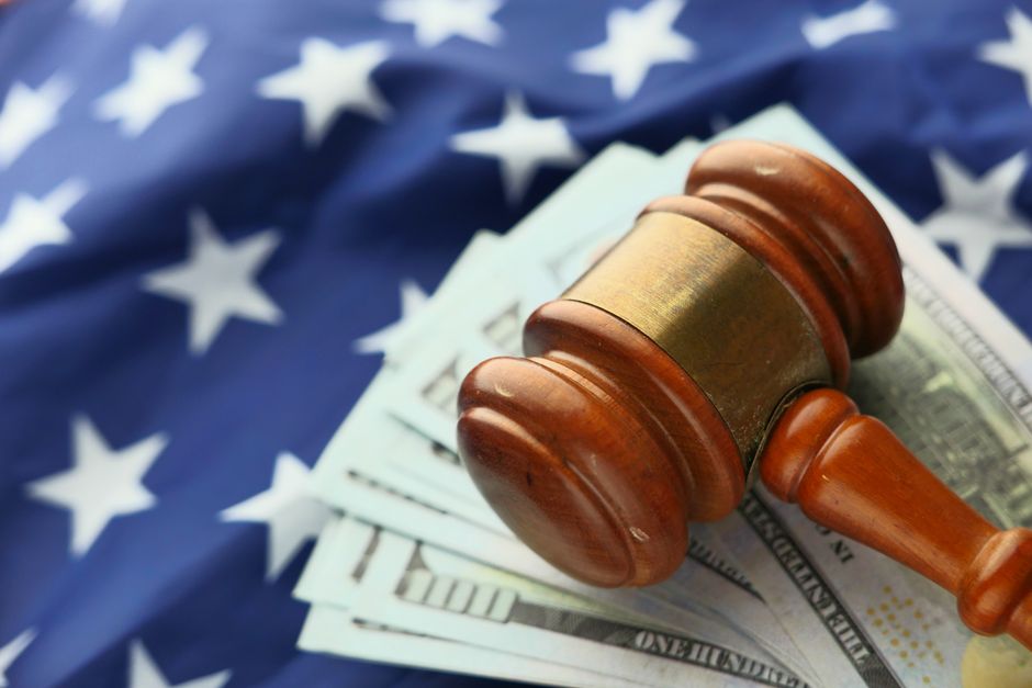 A close view of a wooden judge's gavel resting on a fan of paper banknotes, with a flag hanging out of focus behind it.