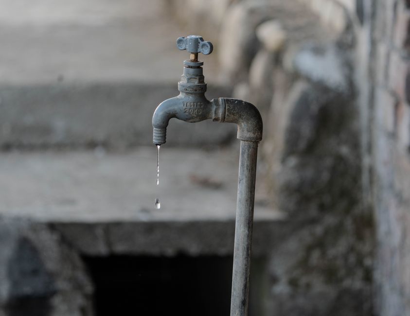 A weathered outdoor metal tap against a rough grey wall, with a single drop of water hanging from the spout.