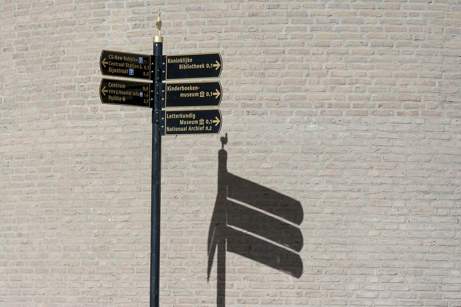 A black and gold directional street sign with several arms pointing different ways, casting a hard shadow on a brick wall in bright sun.